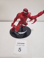 Front angle view of the red Venom toy figure on its black round base with visible 'VENOM' text and logo.