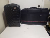 Front view of black hard-shell suitcase and black soft packing bag side by side on a white surface.