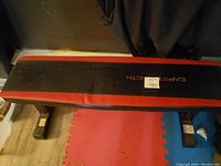 Top down view showing the full length of the workout bench with its black padded cushion bordered by red, placed on a red and blue foam mat.