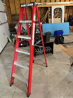 Full view of red 6-foot folding step ladder standing open on a garage floor with wood and other items in the background.