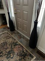 Photo shows two tall black decorative vases positioned on each side of a door, placed on tile flooring next to a patterned area rug.