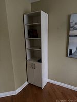 Tall white bookcase with three adjustable shelves and closed lower cabinet with grey pull handles against a beige wall corner.