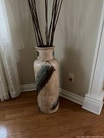 Tall decorative vase with neutral color base, blue and brown accent patches, and thin rope around neck, standing on hardwood floor near wall.