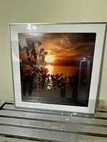 Framed photo art depicting a vibrant sunset over water with silhouetted foliage in foreground, thin silver frame and white matting.