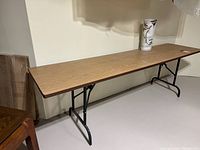 Full side view of the rectangular long table with wood grain laminate top and folding black metal legs in an indoor setting.