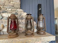 Three weathered metal oil lamps with glass chimneys, one red and two clear, standing on a stone surface.
