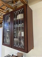 Upper dark wood kitchen cabinet with two leaded glass doors, showing interior shelves with dishes and decorative trim on cabinet edges