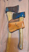 Two axes lying on a pink cloth; one axe has a leather sheath with 'Made in Czechoslovakia' stamped inside, both with wooden handles and worn blue paint on the blade, showing signs of rust and wear.