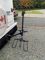 Black metal hitch-mounted bike rack attached to vehicle with two upright hooks padded with foam, horizontal support arms and wheel holders, placed outside on pavement near vehicles.