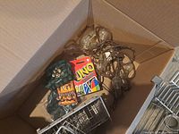 Box containing string lights, UNO cards, green plaid beer cozy, and small storage shelves