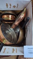 Box of assorted copper cookware showing various pots and pans piled together, showing brass and black handles and varying interior linings