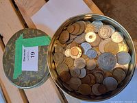 Top view of metal tin container filled with assorted world coins, showing coins in various metals and colors including silver, bronze, and copper tones.
