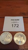 Two silver coins side by side on a wooden surface with a MaxSold label showing lot number 172.