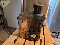 Electric juicer assembled with black base, dial control, clear juice container attached, and power cord visible on wooden surface.
