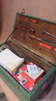 Open green wood tool chest showing older hand tools mounted on the lid, papers and boxed items inside the chest.