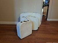 Three soft sided suitcases standing upright on wood floor, light beige color, showing front and top handles of each.