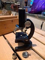 Side view showing black metal microscope body with chrome column, adjustable focus and stage with clips, resting on a wooden surface with original box in background.