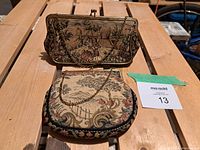 Two vintage needlepoint tapestry evening bags on a wooden surface. One bag is rectangular with a clasp frame showing a detailed scenic needlepoint design with figures and greenery. The other is rounded with a landscape needlepoint pattern and metal chain strap.