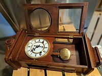 Open clock showing white dial with Roman numerals and brass pendulum inside wooden case with glass panels.