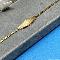 Close-up view of delicate gold ID bracelet with patterned edge plate and fine chain.