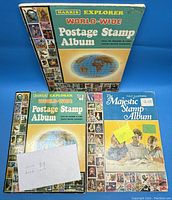 Three stamp albums including two Harris Explorer World-Wide Postage Stamp Albums and one Majestic Fully Illustrated Stamp Album positioned on blue background with a note indicating loose and album stamp counts.