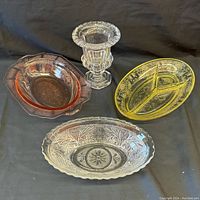 Photo showing three pieces of Depression glass in different colors - yellow divided dish, pinkish-red bowl, clear oval bowl - and one clear glass vase with pedestal base, all on black background.