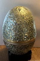 Image showing full view of the large egg-shaped brass box with detailed enamelled floral designs, resting on a black stand placeholder.