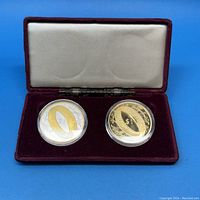 Both coins in protective capsules inside the red plush case with beige lining on the lid.