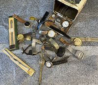 Photo showing several old wristwatches with varied metal and leather bands scattered near a rusted tin container.