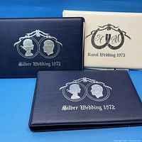 Three stamp albums: two blue Silver Wedding 1972 albums and one white Royal Wedding 1973 album, arranged side by side on blue background.