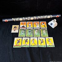 Full view of 15 assorted baseball cards laid out with the rolled Toronto Blue Jays poster and white metal bell with insignias.