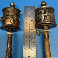 Two Tibetan prayer wheels shown side by side with a ruler indicating approximate height around 7 inches, details of the copper/brass tonal metal drum and wooden handle with wire wrapping visible.
