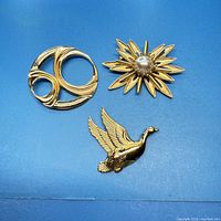 Three gold tone vintage brooches displayed on a blue background: abstract circular, flower with pearl center, and flying bird.