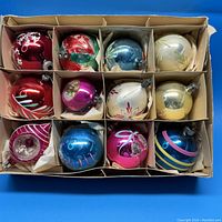 Top-down view showing all 12 vintage glass Christmas ornaments packed in divided cardboard box with tissue paper protection. Various colors and designs visible.