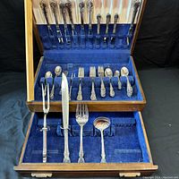 Full open view of wooden box with multiple slots for flatware. Items arranged in the top lid slots and main compartment with blue velvet lining. Includes forks, spoons, and serving utensils.