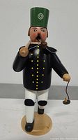 Front view of the Erzgebirgische Miner wooden smoker figurine, showing black jacket with gold dots, white pants, green hat, and pipe in mouth.