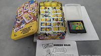 Lot of The Simpsons domino set with metal tin, domino tiles, and instructions sheet beside Sega Game Gear cartridge in translucent case.