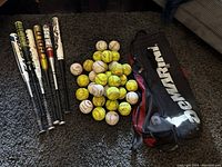 Photo showing six baseball/softball bats standing vertically with grips visible, multiple yellow softballs and white baseballs grouped on carpet, and a black/red DeMarini bag.
