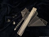 Photo shows six items: a pair of cuff links, a ring, a tie tack, a bracelet, a necklace, and its packaging. Items are black and silver-tone with fleur-de-lis and geometric designs.