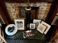 Photo showing multiple framed artworks, a round white mirror, new wind chime with tags hanging, and floral patterned chair cover on floor in fireplace opening.