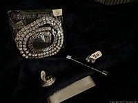 Photo showing a men's bling jewelry set including a 24-inch rhinestone tennis chain necklace in packaging, a silver-tone ring, and a matching pin or small earrings, all on black fabric.