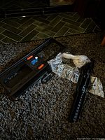 Black metal TV wall mount bracket alongside multiple sealed bags of hardware including screws, bolts, and wall anchors laid out on carpet.