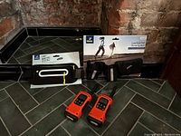 Two black running belts, one in original packaging, and two orange and black walkie talkies displayed on floor against brick wall.