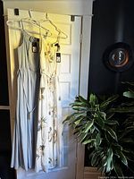 Two dresses hanging on hangers on a white door. One is light blue-gray sleeveless maxi and the other is white floral print maxi with long sleeves and waist tie.