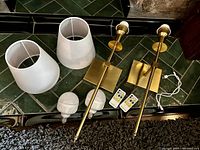 Photo showing two wall mount sconce bases in brass-tone finish, two white fabric lampshades, two rechargeable LED bulbs, and two white remote controls with multi-button layouts. Items arranged on green tiled floor near a brick wall.