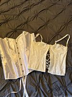 Three white corsets laid on a dark tufted fabric: one satin corset with front closures and ribbons, two lace corsets with straps and lace-up details.