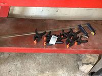 Set of black plastic hand spring clamps with orange gripping tips and one large blue-handled 32 inch F clamp on a red surface.