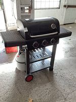 Front angle view of the black Coleman propane barbecue grill showing three red knob controls, lid with temperature gauge, side shelves, and propane tank attached below.