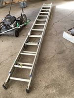 Full view of aluminum extension ladder laid on concrete floor next to a lawn mower, showing total length and condition.
