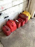 Six fuel cans lined up on a concrete floor against a wall. Five red cans and one yellow can. Various sizes and some with caps attached on the top. Shows overall condition and color.